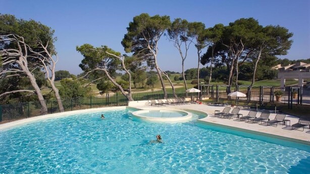 Una piscina con una sección circular, rodeada de sillas de descanso y sombrillas, con árboles y un campo de golf al fondo.