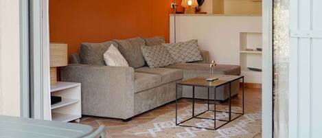 Una sala de estar con un sofá gris, una mesita de centro y una lámpara. Hay una zona de cocina con armarios y una lámpara colgante.