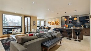 Una sala de estar moderna con un amplio sofá modular, un banco y un comedor, con una cocina al fondo.