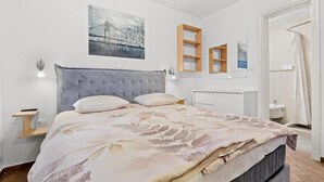 A bedroom with a bed, bedside tables, a headboard, a painting, and a bathroom with a sink and shower.