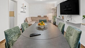 A modern dining area with a long table, chairs, a bowl of fruit, a remote control, and a television on the wall.