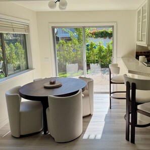 Un espace repas moderne avec une table ronde et des chaises rembourrées, une cuisine avec des meubles blancs et une vue sur un jardin grâce à de grandes portes coulissantes.