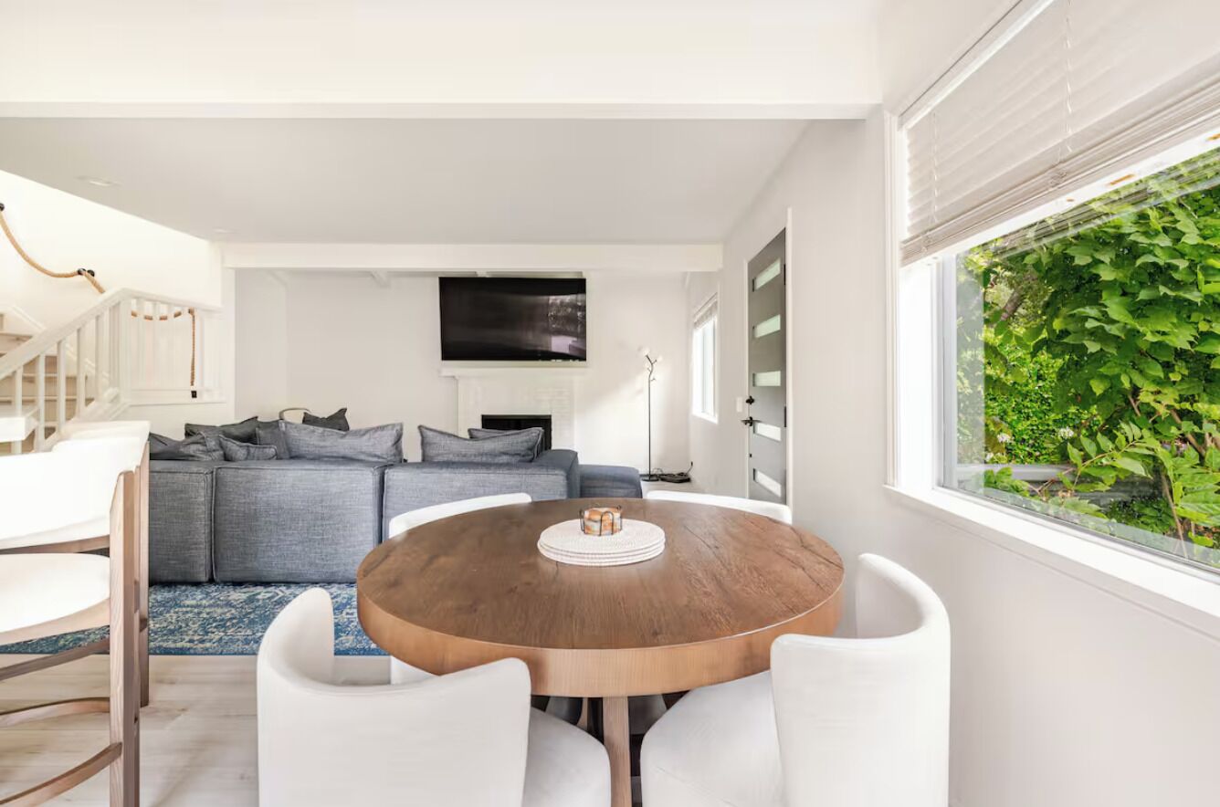 A modern living room with a grey sofa, a round wooden dining table with white chairs, and a large window with a view of greenery.