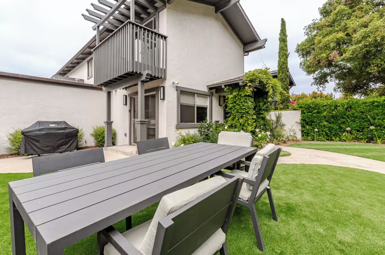 A modern house with a patio area featuring a dining table and chairs, a covered grill, and a small outdoor kitchen.