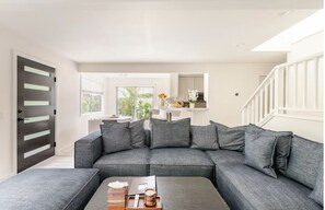 En moderne stue med en grå hjørnesofa, et træbord og udsigt til køkken og have gennem store vinduer.