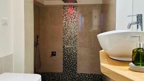 Een badkamer met een witte badkuip, een douche met een wand van mozaïektegels, en een houten wastafelmeubel met een groene zeepdispenser.