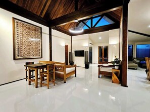 A spacious room with a high ceiling, wooden beams, a large window, a dining table with chairs, a sofa, and a television.