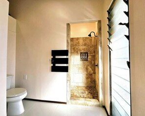 A bathroom with a toilet, a shower area with a tiled wall, and a door leading to another room. - Breathtaking Oceanfront Home on Lava Rock Beach. (Pahoa)