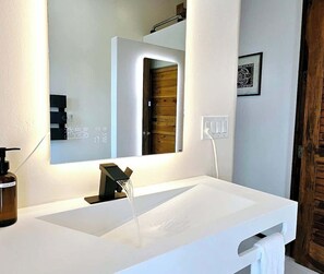 A modern bathroom with a white sink, a large mirror, and a wooden door.