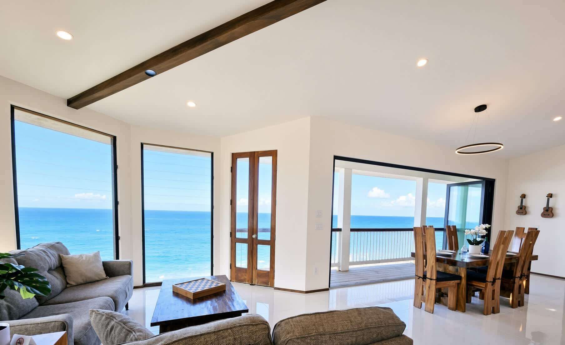 A spacious living area with large windows, a wooden ceiling beam, and a dining area with a view of the sea.