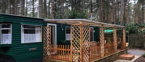 A green mobile home with a fenced porch area, surrounded by a forest.