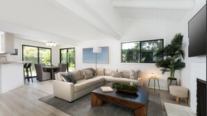 A spacious living room with a large L-shaped sectional sofa, a coffee table, and a dining area with a bar. There is a large window providing natural light and a view of greenery outside.
