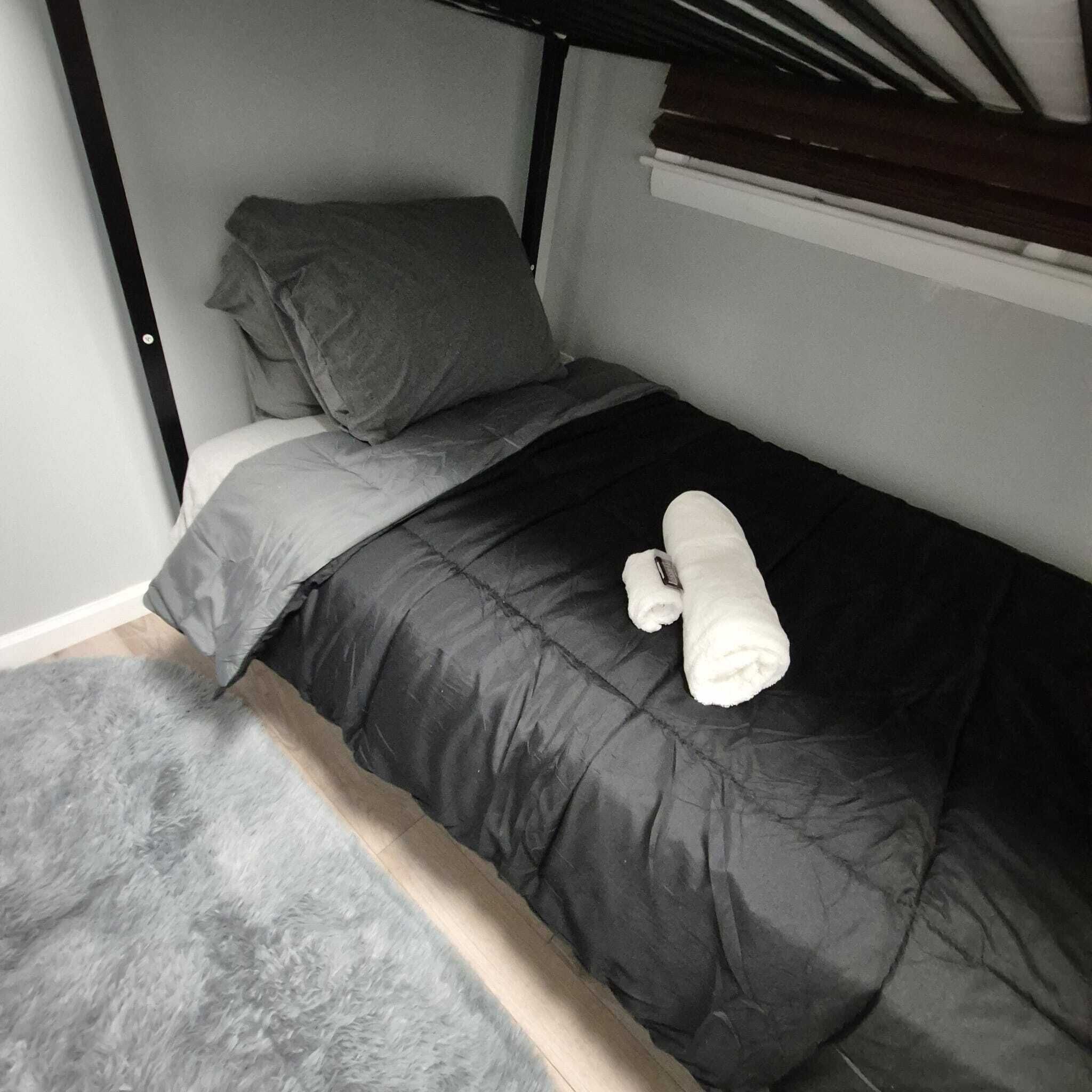 Een stapelbed met een grijs matras en kussens, een opgerolde handdoek en een pluizig grijs vloerkleed.