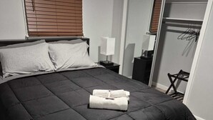 A bedroom with a black bedspread, a grey bench, a wooden headboard, a bedside table with a lamp, and a closet with hangers.