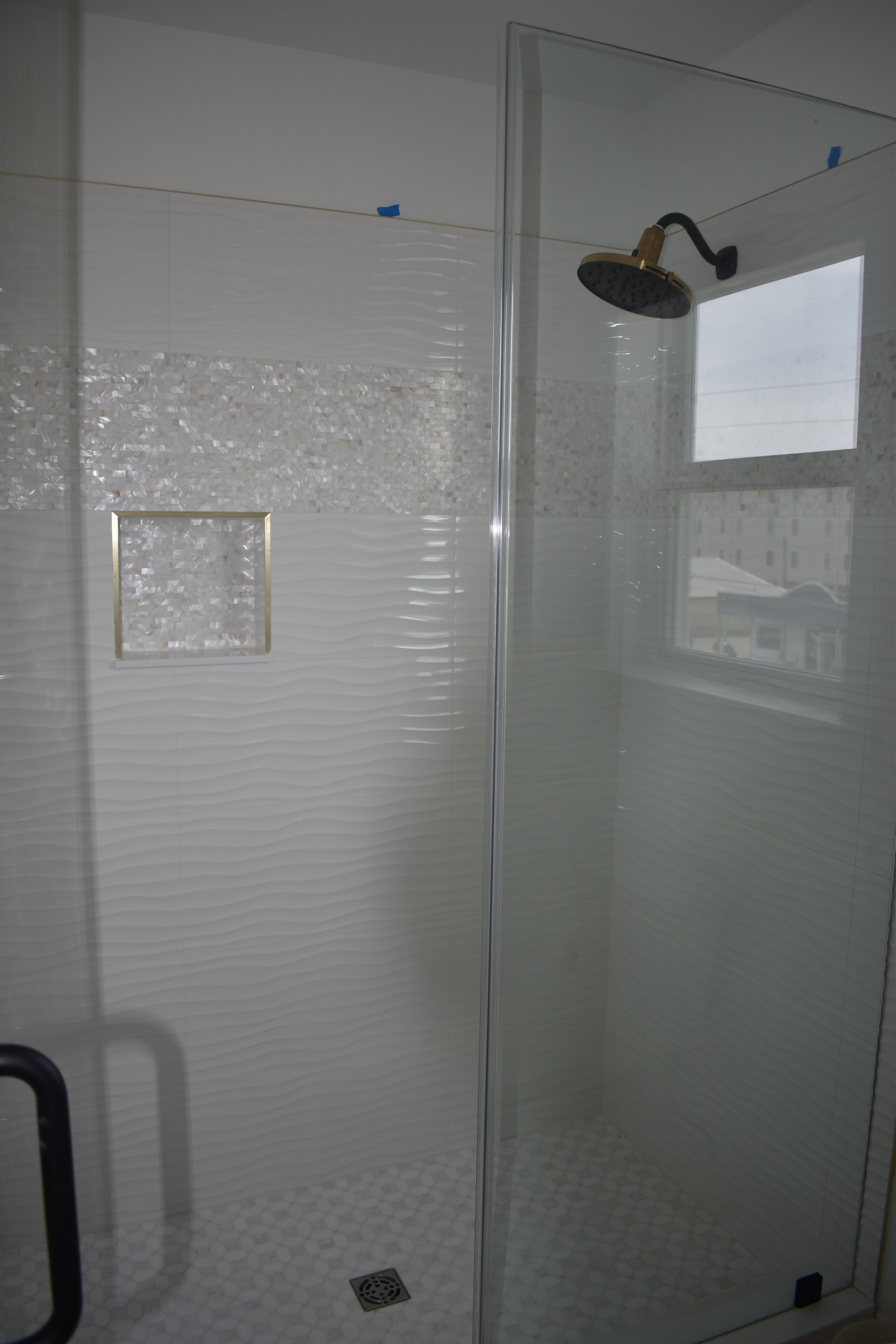 Une douche fermée par des parois en verre, dotée d’un pommeau de douche à main, d’une petite fenêtre et d’un mur à texture.