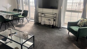 Ein möblierter Raum mit einem Glastisch, einem an der Wand montierten Fernseher, einem Esstisch mit Stühlen und einem Sofa mit Kissen.