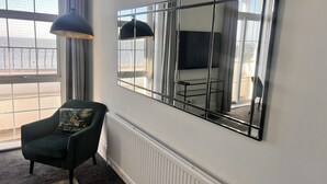 Ein Zimmer mit großem Fenster, einem grünen Sessel, einer schwarzen Pendelleuchte und einem großen Spiegel an der Wand.