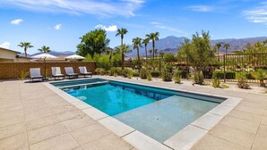 Una piscina rectangular con un borde de azulejos, rodeada de sillas de descanso y palmeras, con montañas al fondo.
