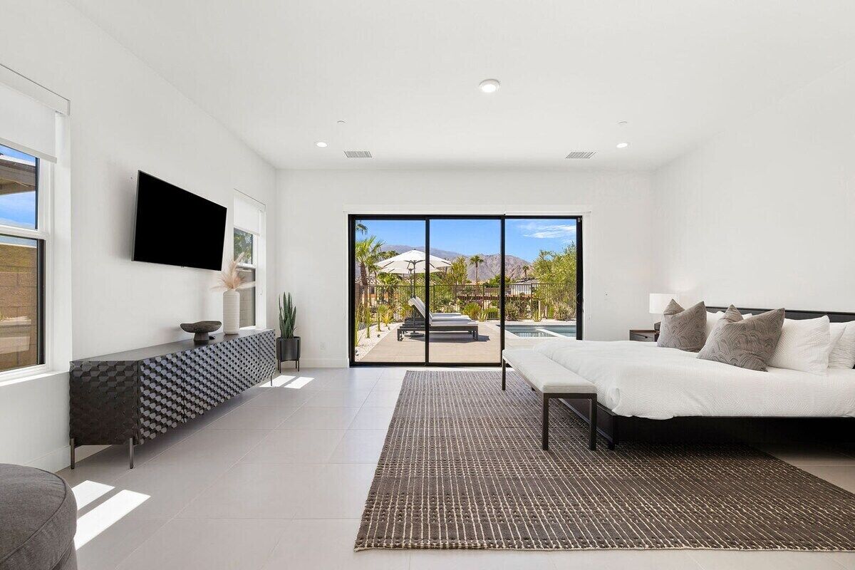 A modern bedroom with a bed, a TV, a console table, and a view of a pool and mountains.