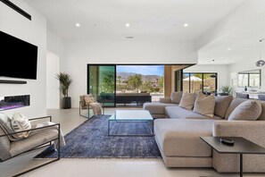 Una sala de estar moderna con un televisor de pantalla plana grande, un sofá modular, una mesa de centro de vidrio y vistas a un paisaje montañoso a través de grandes ventanales.