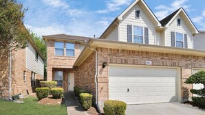 A two-story house with a brick facade, a white garage door, and a well-maintained lawn with trimmed bushes.