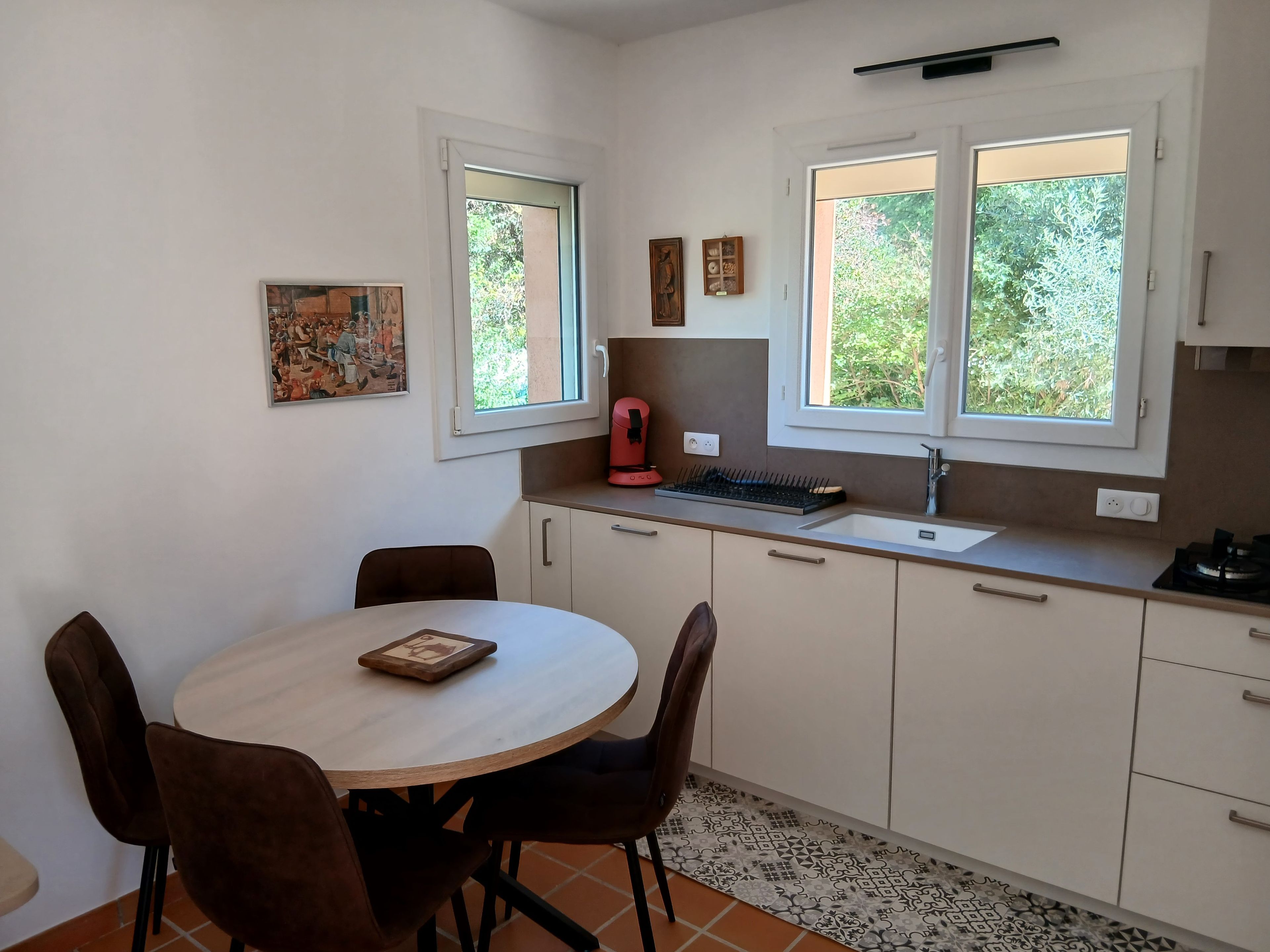 Une cuisine compacte avec un coin repas, comprenant une table ronde, des chaises, une machine à café et deux fenêtres qui laissent entrer la lumière naturelle.