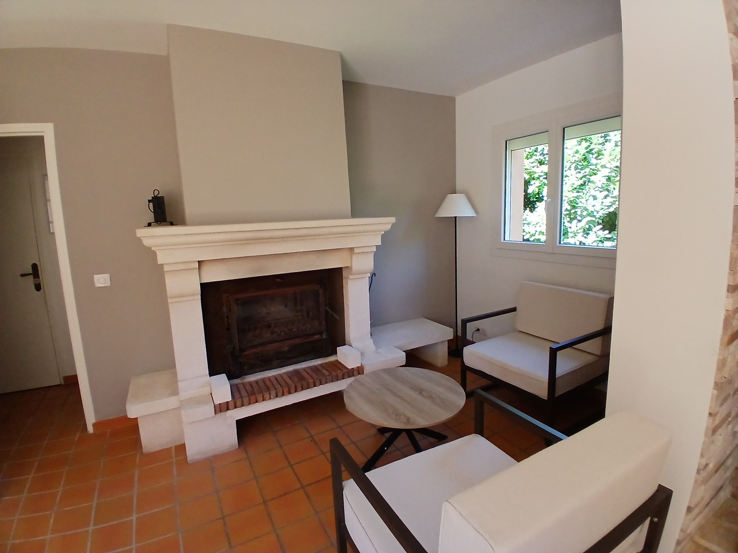 A room with a fireplace, a window with a view of greenery, a lamp, and a seating area with a chair and a table.