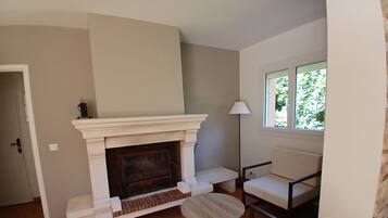 A room with a fireplace, a window with a view of greenery, a lamp, and a seating area with a chair and a table.