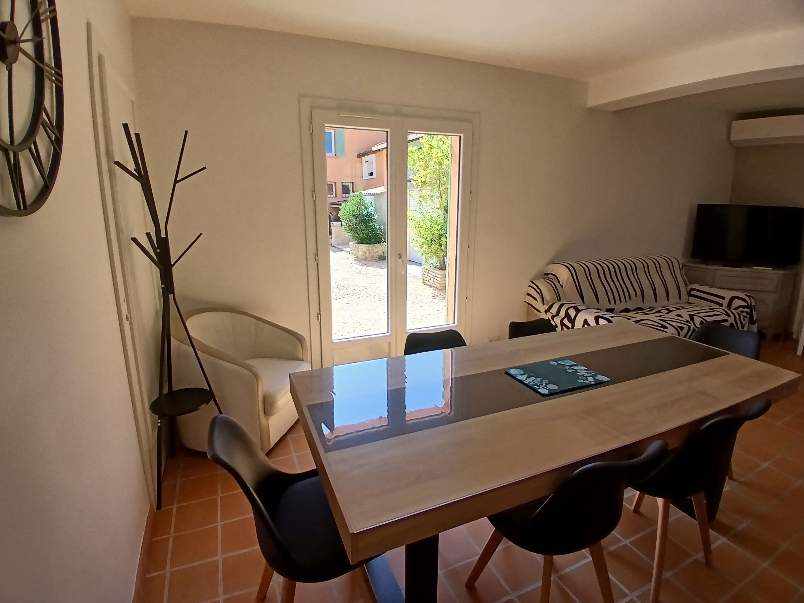 A dining area with a table and chairs, a sofa, a television, and a view through a door to the outside.