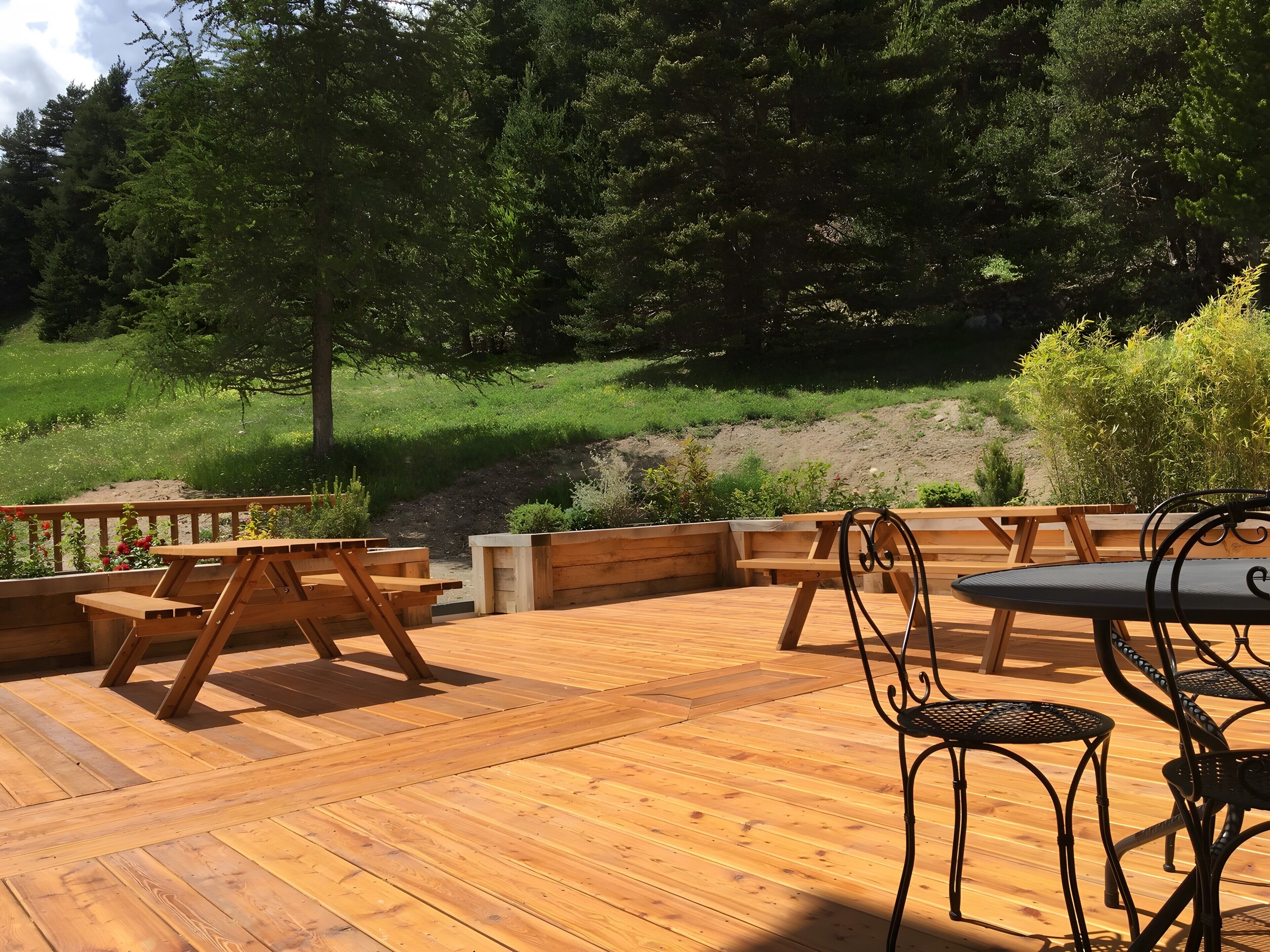 Una terraza de madera con mesas y sillas de picnic, rodeada de follaje y árboles.