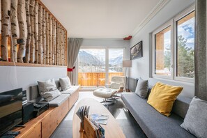 En moderne stue med sofa, lænestol og sofabord, en trævægsudsmykning og et stort vindue med udsigt til et bjerglandskab.