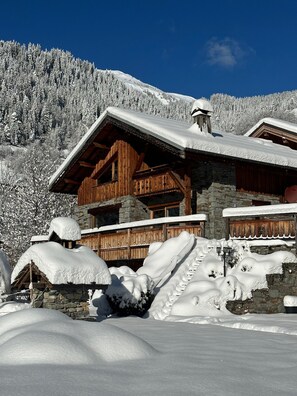 A wooden chalet with a stone foundation, surrounded by snow-covered trees and a clear blue sky.