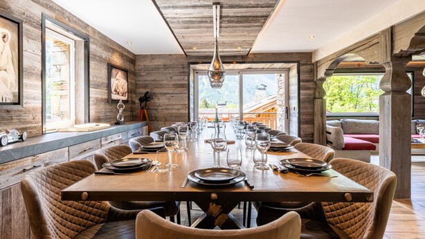 Une longue table à manger dressée pour un repas, des murs en bois et de grandes fenêtres.