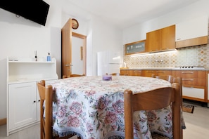 Ein Essbereich mit rundem Tisch, Stühlen, einem an der Wand befestigten Fernseher sowie einer Küche mit Holzausstattung und gefliestem Rücksprung.