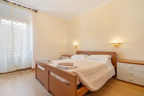 A bedroom with a wooden bed, bedside tables, a nightstand, and a window with sheer curtains.