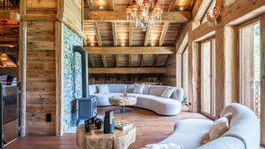 L’intérieur d’une cabane en rondins, avec un mur en pierre, des poutres en bois au plafond, une cheminée, un grand canapé blanc et un lustre.