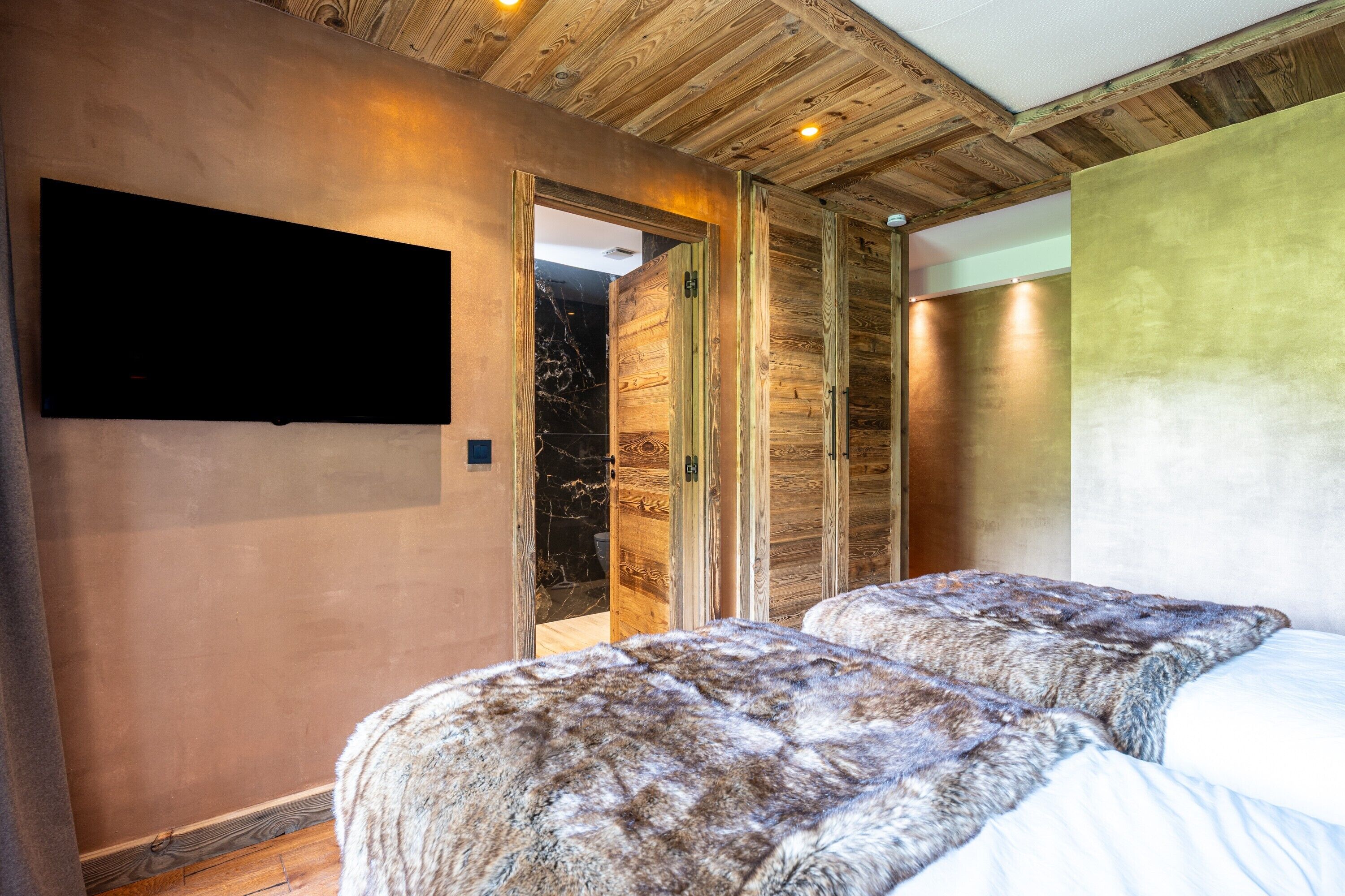 A room with a wooden ceiling, a flat-screen TV mounted on the wall, and a bed with a fur throw.