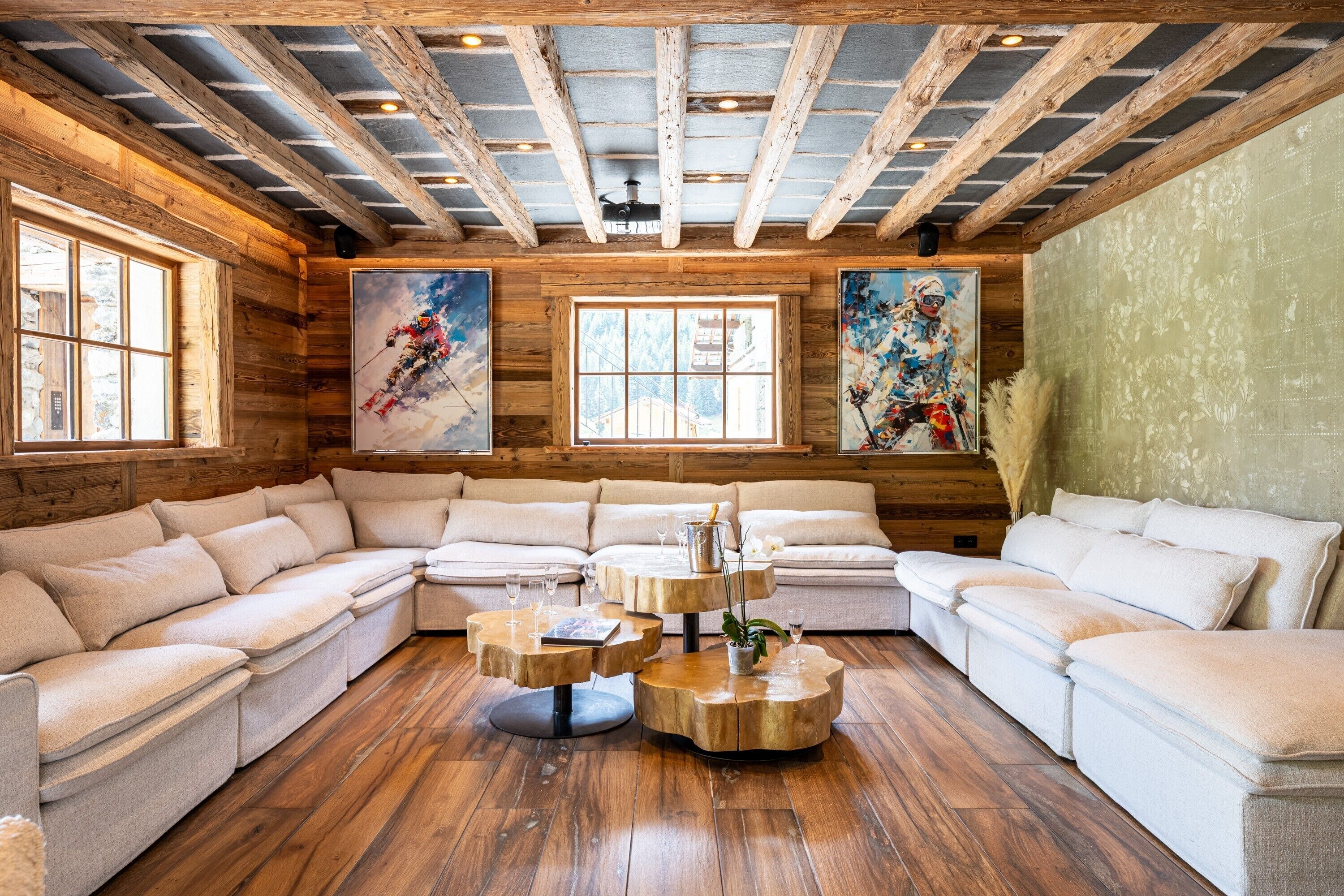 A log cabin interior with a large sectional sofa, wooden coffee tables, and two large windows with paintings.