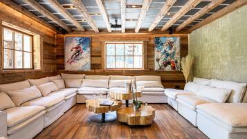A log cabin interior with a large sectional sofa, wooden coffee tables, and two large windows with paintings.