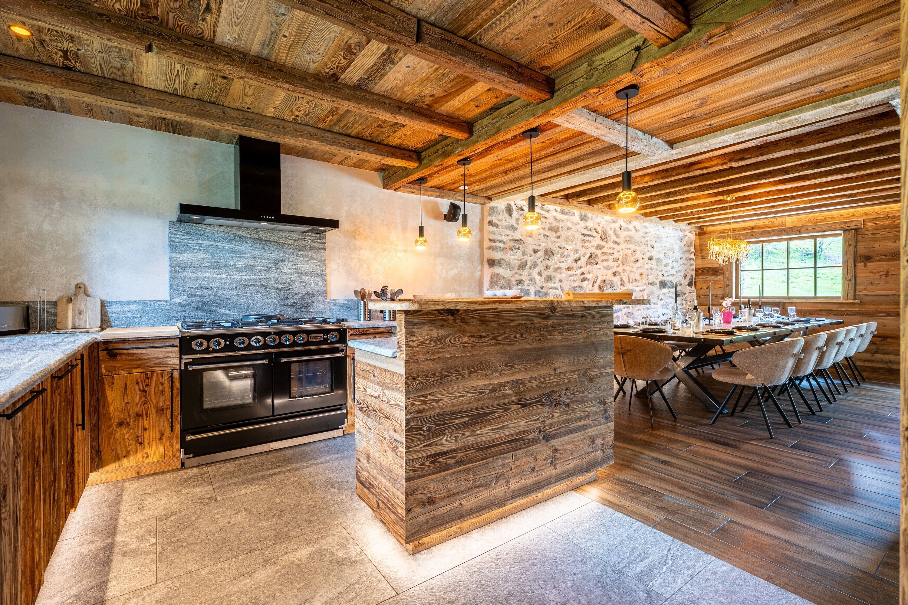 A rustic kitchen with a large island, wooden beams, stone wall, and a dining area with chairs.