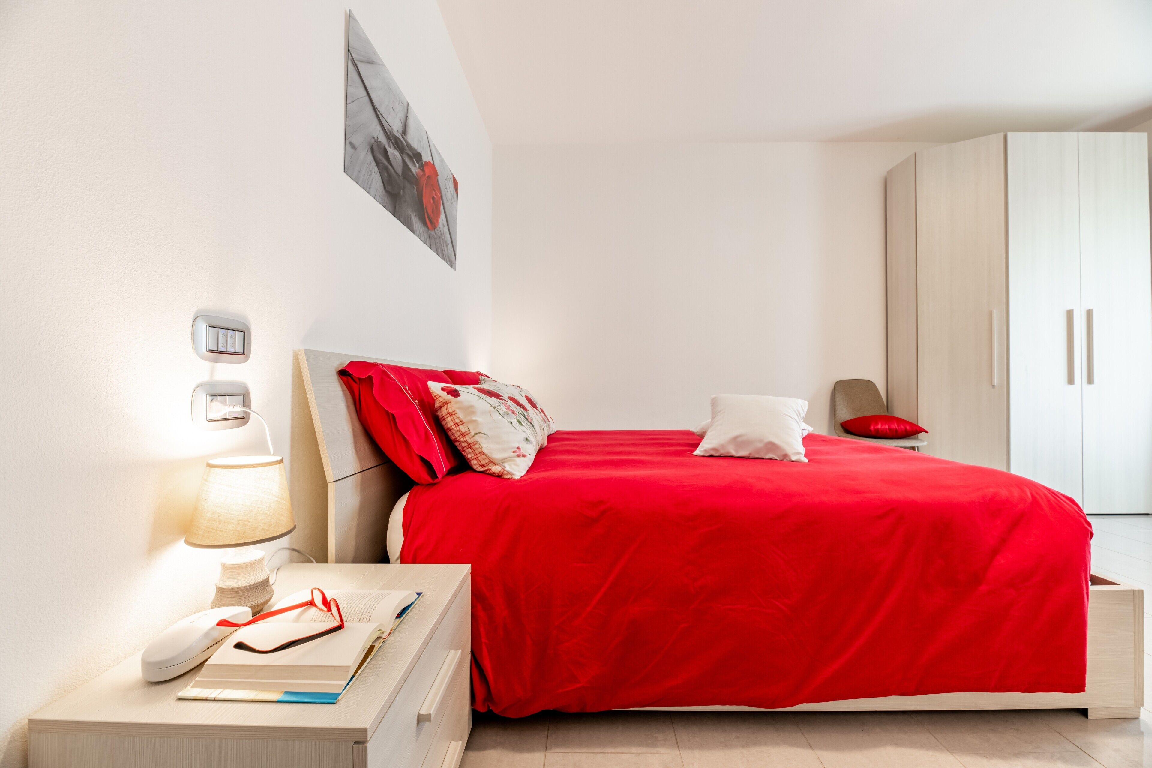 Une chambre avec une couvre-lit rouge, une lampe de chevet, une table de chevet avec un livre et des lunettes, un thermostat fixé au mur et une armoire.