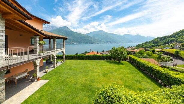 Een huis met een balkon dat uitkijkt over een meer, stenen zuilen en een verzorgd gazon.