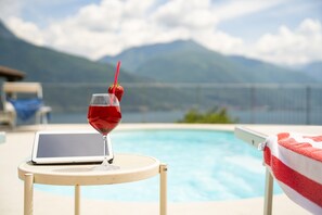 Une scène de bord de piscine : un ordinateur portable posé sur une chaise, un verre d’une boisson rouge avec une paille, et une serviette sur une autre chaise.