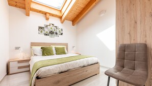 Een slaapkamer met dakraam, houten balken in het plafond, een bed met groene beddengoed, een nachtkastje en een stoel.