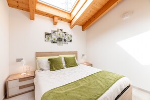 Une chambre avec un puits de lumière, un lit avec des coussins verts, une table de chevet avec une lampe et une œuvre d’art murale.