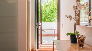 Una puerta corredera abierta que da a un balcón con vistas a zonas verdes, una planta en maceta y un recipiente de vidrio sobre una mesa de madera.