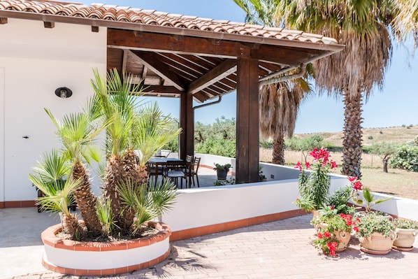 A covered patio area with a table and chairs, surrounded by potted plants and palm trees.