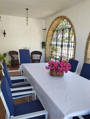 Un coin repas avec une table blanche, des chaises bleues et un panier en osier contenant des fleurs roses.