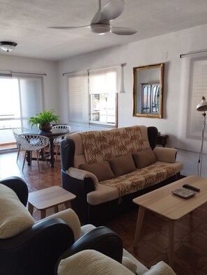 Un salon comprenant un canapé, des fauteuils, une petite table, un ventilateur de plafond et une fenêtre avec des stores.
