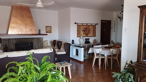 A living room with a fireplace, a ceiling fan, a dining area with a table and chairs, and a kitchen area with a fruit bowl. - Chalet El Paraíso with sea views, private terrace, and Wi-Fi (Laguna del Portil/Huelva)